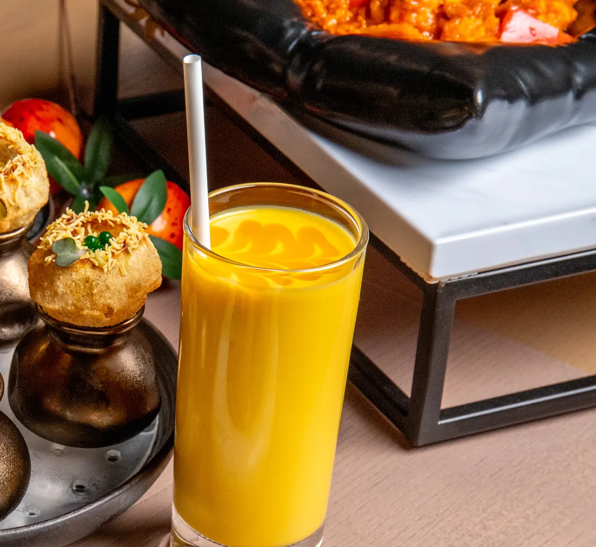A tall glass of mango lassi with a paper straw, beside pani puri on small bronze pots