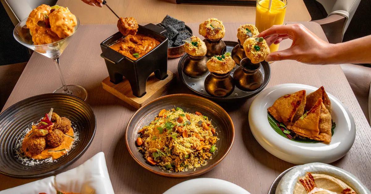 A hand reaching for pani puri on a table of small plates and samosas