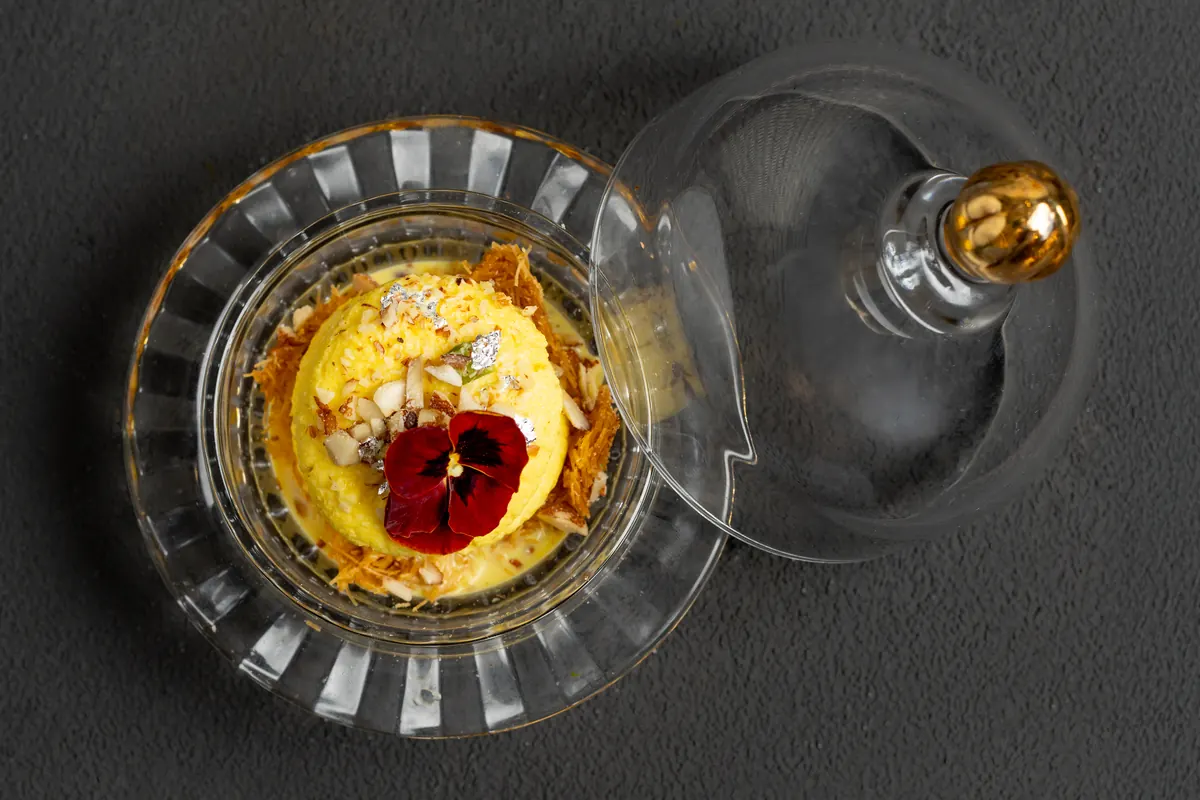 Ras malai with saffron foam and an edible flower in a glass dish, beside its glass cloche