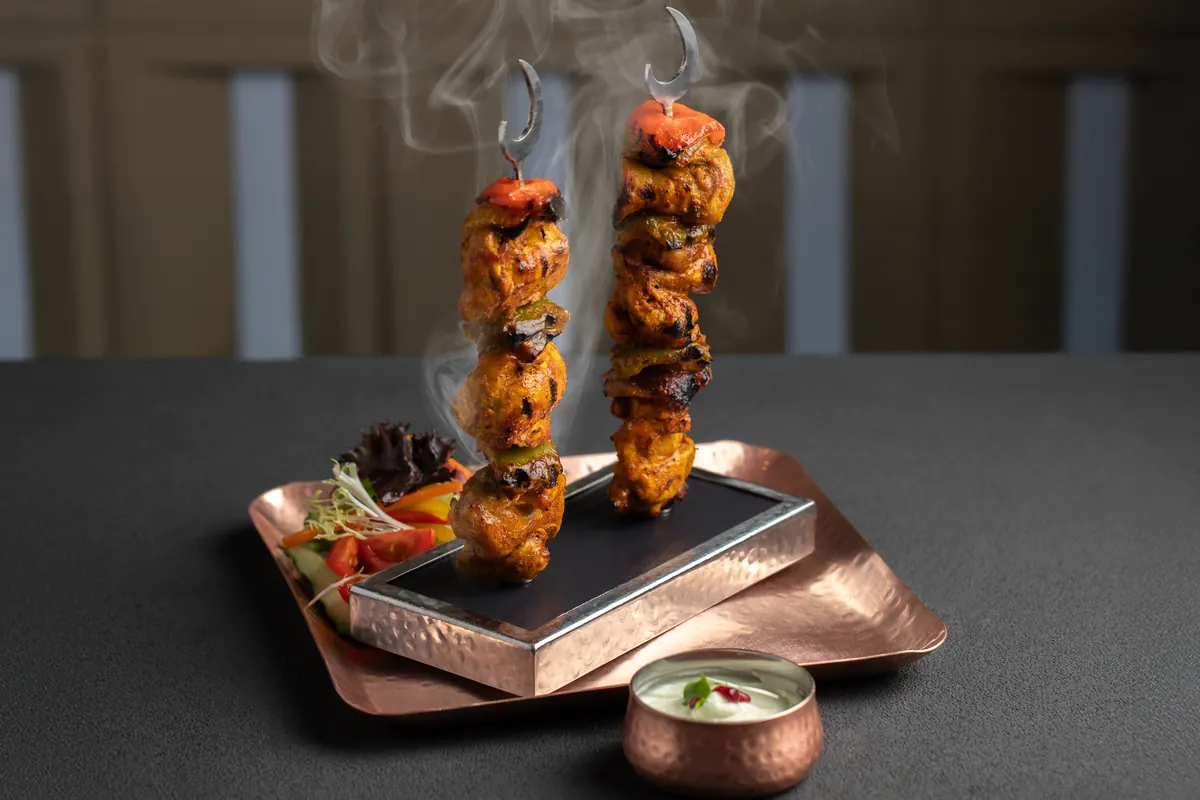 Two skewers of shish touk standing on a hot stone, with salad and a small copper pot of dip